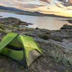 A photo of a camping tent set up by a body of water with the sunset in the background.