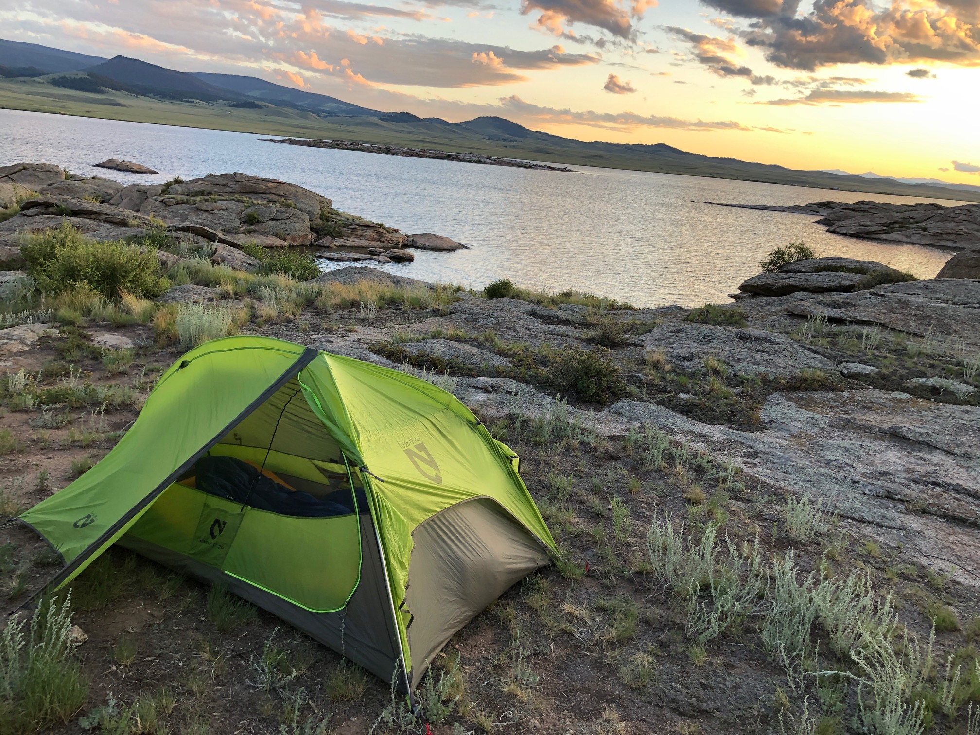 A photo of a camping tent set up by a body of water with the sunset in the background.